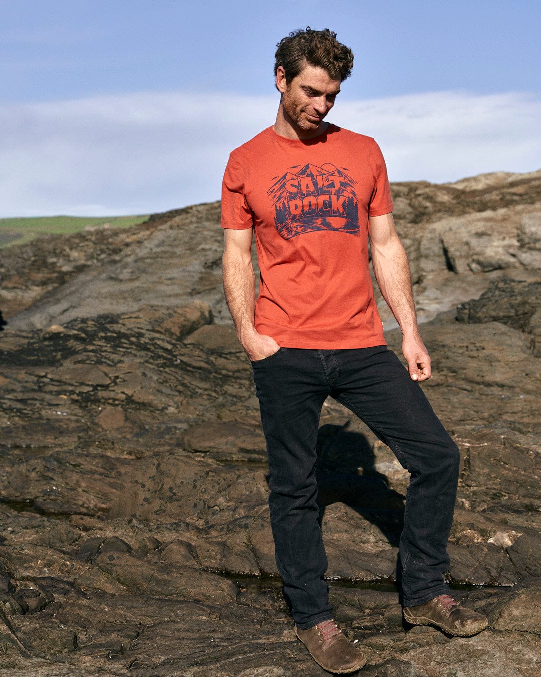 A stylish man in a Saltrock Wood Carve Logo - Mens Short Sleeve T-Shirt - Red standing on rocks in a mountain scene.