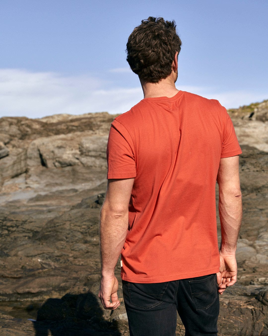 A man wearing a stylish Saltrock Wood Carve Logo - Mens Short Sleeve T-Shirt - Red standing on a rocky beach.