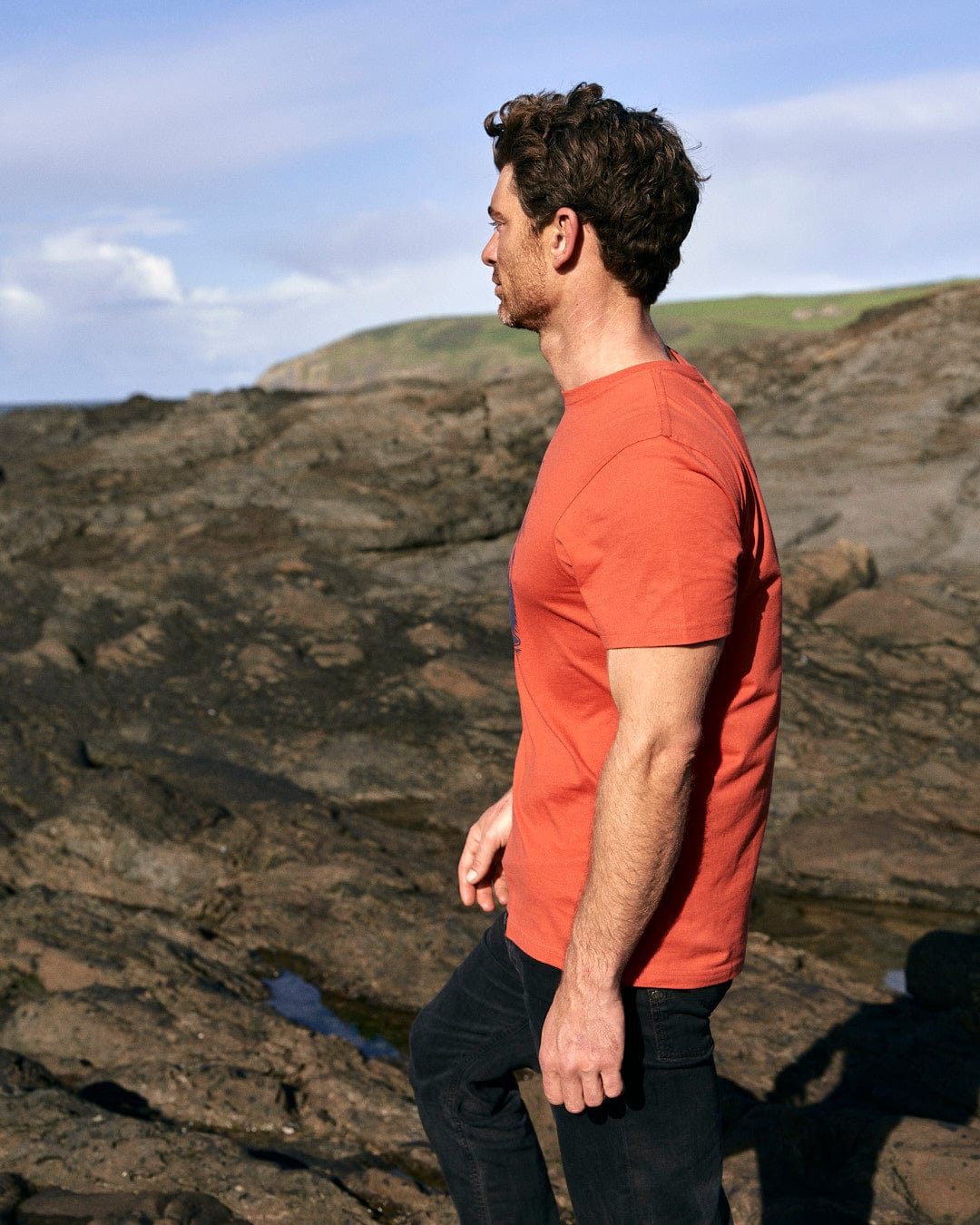 A stylish man wearing a Saltrock Wood Carve Logo - Mens Short Sleeve T-Shirt - Red standing on a rocky beach.