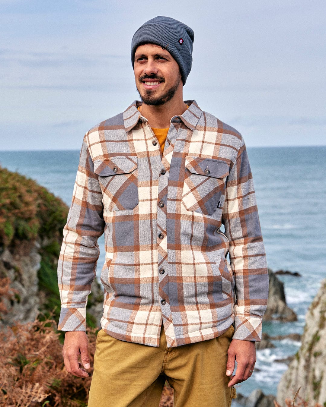 A stylish man wearing a plaid shirt and beanie standing by the ocean, exuding warmth with his Saltrock Woody - Mens Sherpa Lined Shacket - Brown.