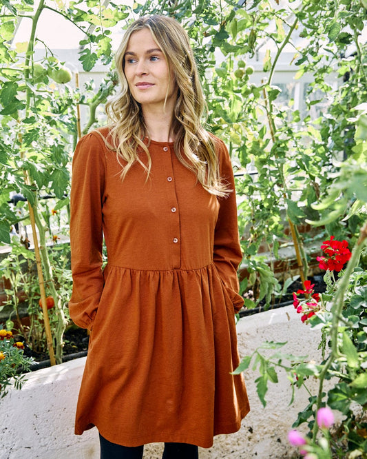In a garden setting, an individual is elegantly adorned in the Saltrock Winnie Women's Dress in dark orange. The rust-colored jersey slub fabric dress features long sleeves that enhance its elasticated waist, offering a flattering silhouette. Surrounding green plants and red flowers add a vibrant touch to this charming scene.