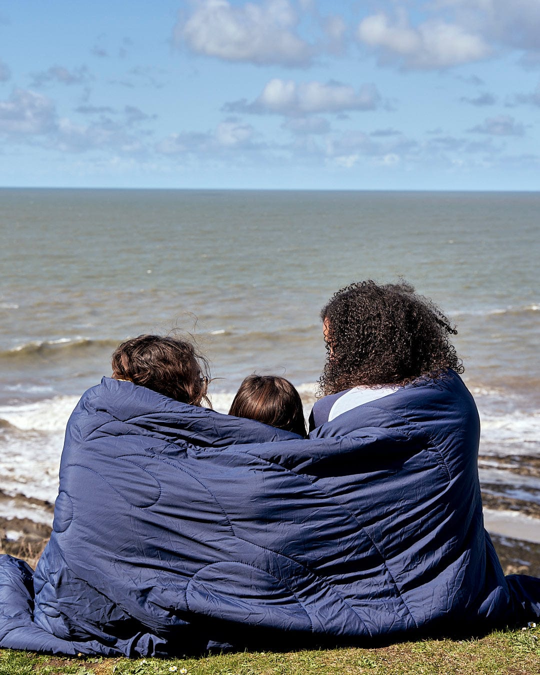 A group of people sitting in a Yogi - Camping Blanket - Blue by Saltrock on a hill overlooking the ocean.