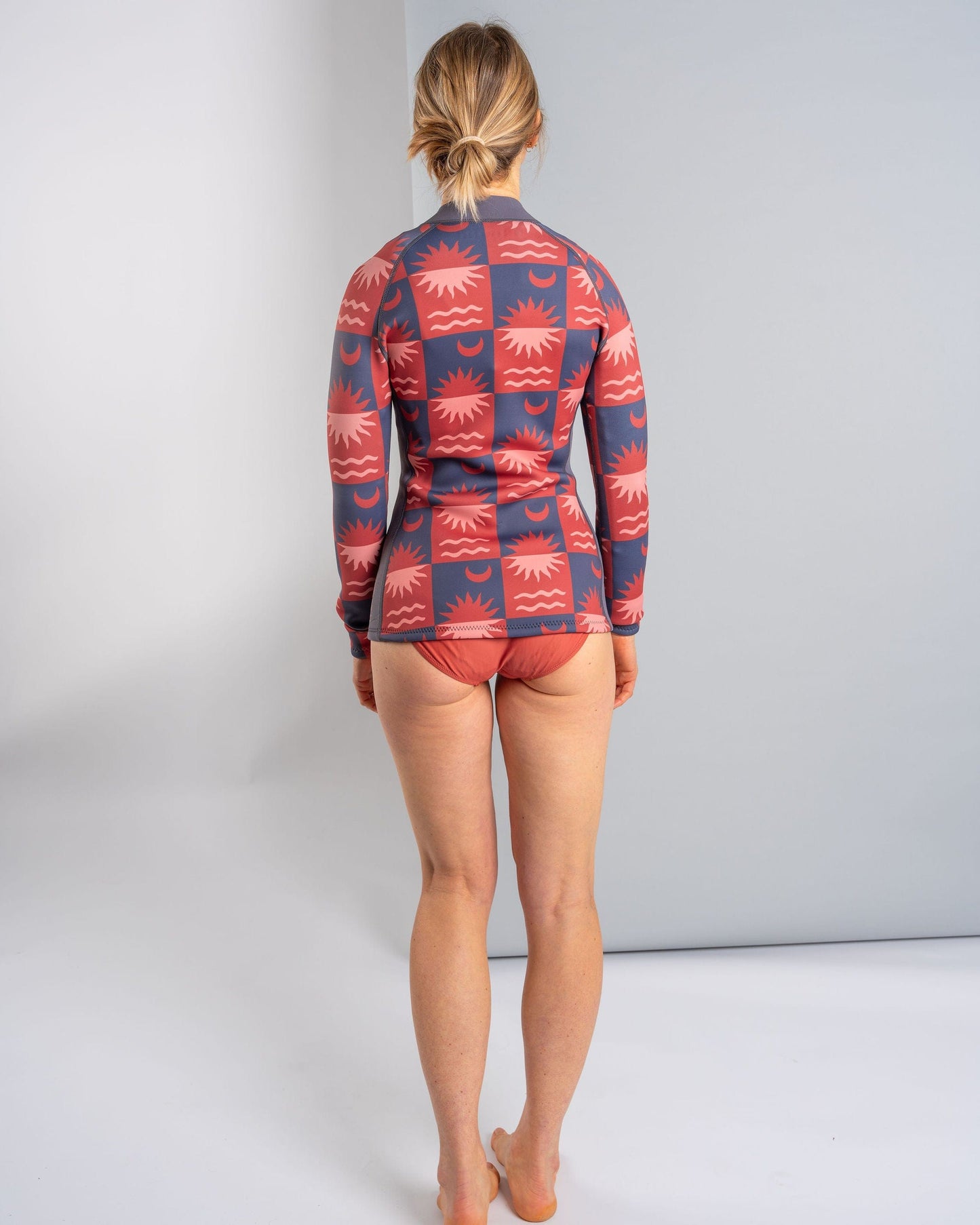A person stands barefoot facing a wall, wearing the Saltrock Equinox Women's Wetsuit Jacket in dark grey/orange with flat locked stitching, paired with matching bikini bottoms.