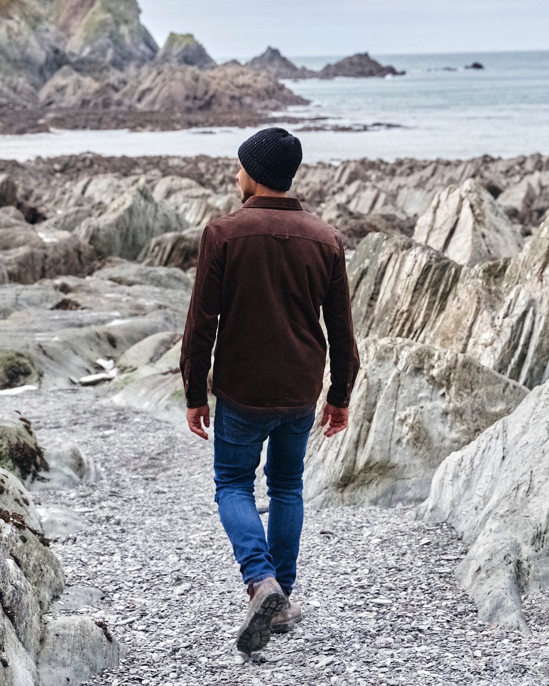 A man strolling along a rugged path close to the ocean, enjoying the comfort of his Zeke - Mens Cord Borg Lined Shacket in Dark Brown by Saltrock.