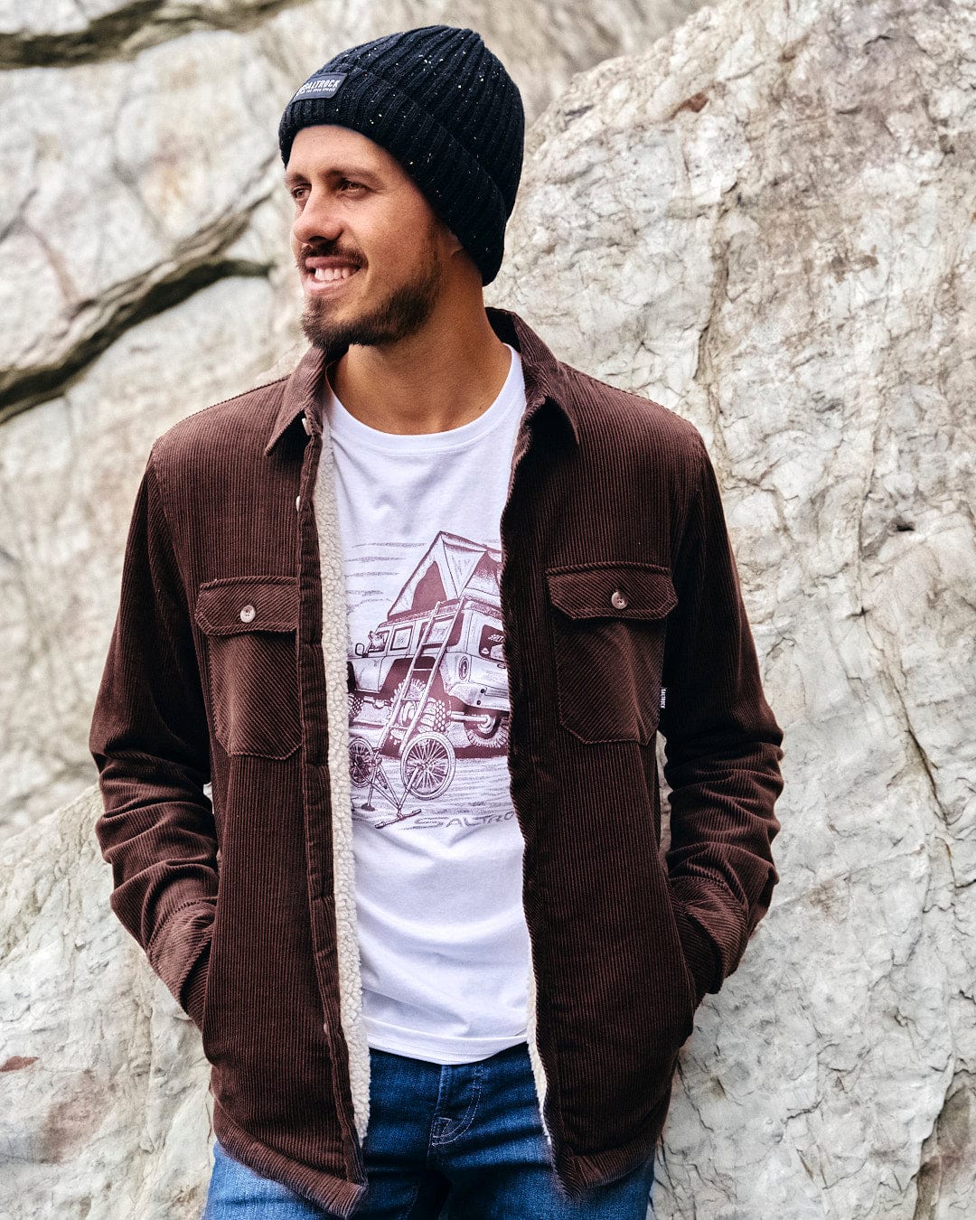 A stylish man in the Saltrock Zeke - Mens Cord Borg Lined Shacket - Dark Brown is comfortably leaning against a rock.
