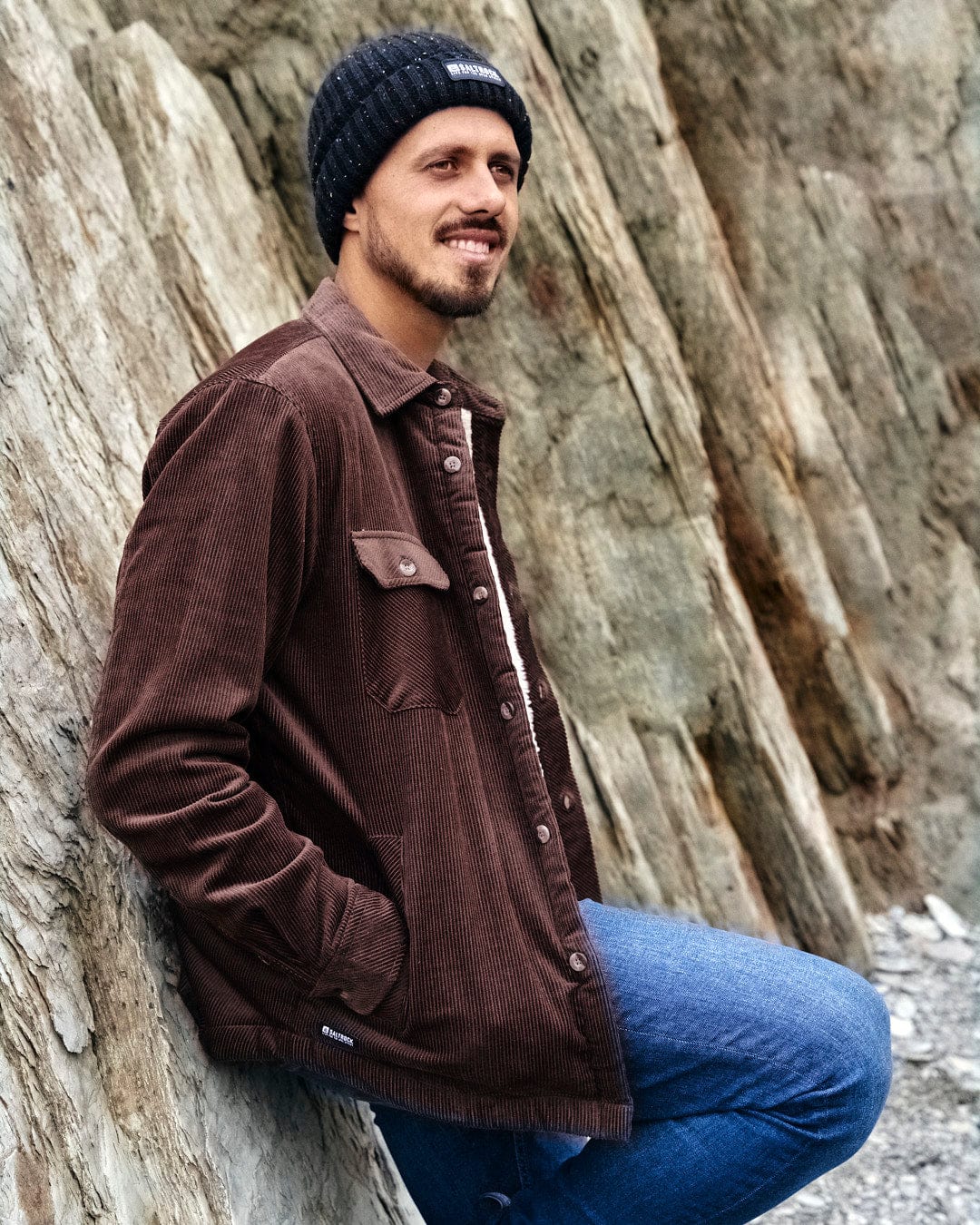 A stylish man sitting on a rock with a Zeke - Mens Cord Borg Lined Shacket - Dark Brown by Saltrock on his head, exuding comfort.