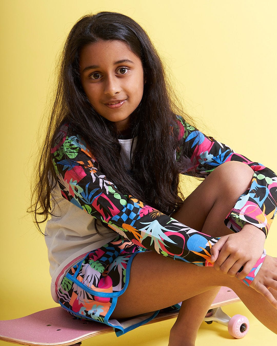 A young girl sitting on a Saltrock Zephyr Tok - Kids Raglan Long Sleeve T-Shirt - White skateboard.