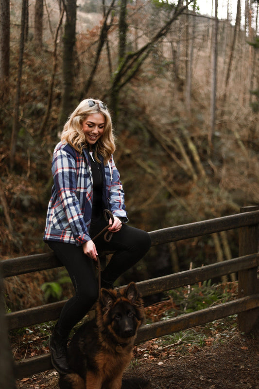 A woman in a Myla - Womens Hooded Checked Shacket - Blue Check from Saltrock, with front pockets and sunglasses smiling while sitting on a wooden fence in a forest, holding a leash with a German shepherd dog next to her.