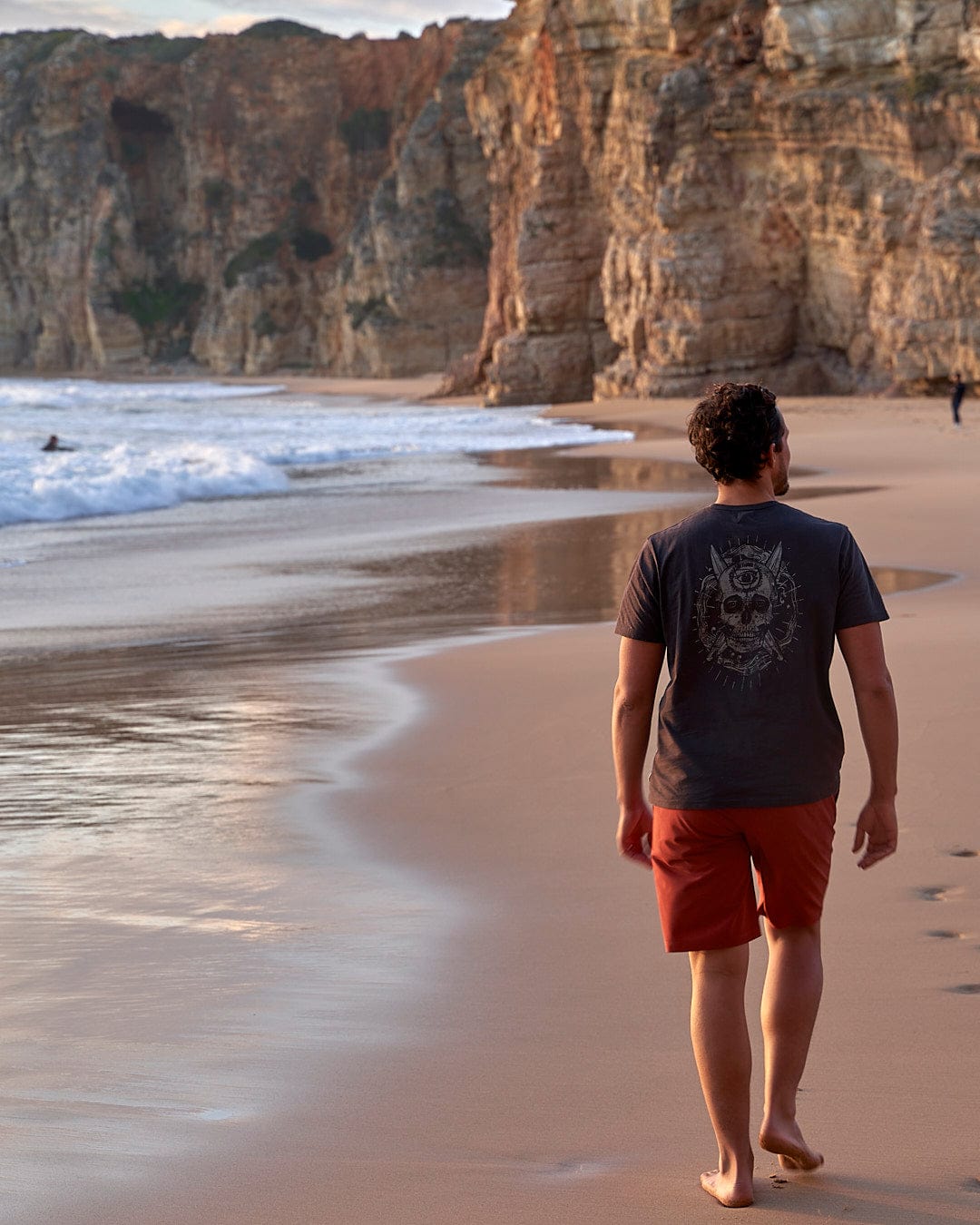 A man walking on a beach near the ocean wearing the Amphibian II - Mens Boardshort - Red by Saltrock.