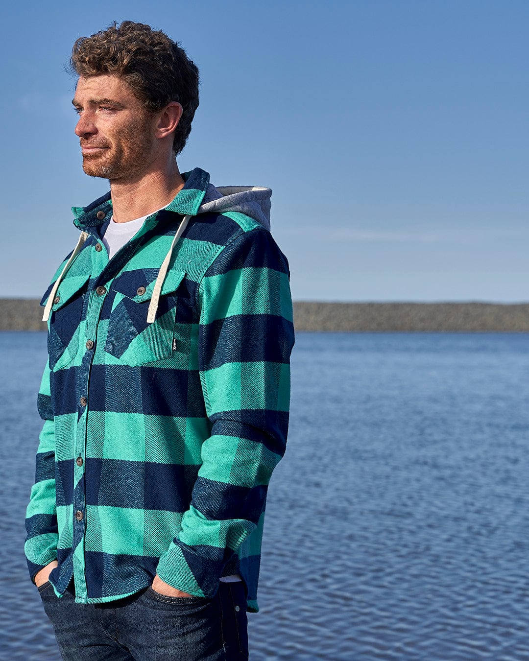A man wearing a Saltrock Beale - Mens Heavy Weight Hooded Shirt - Dark Green standing by the water.
