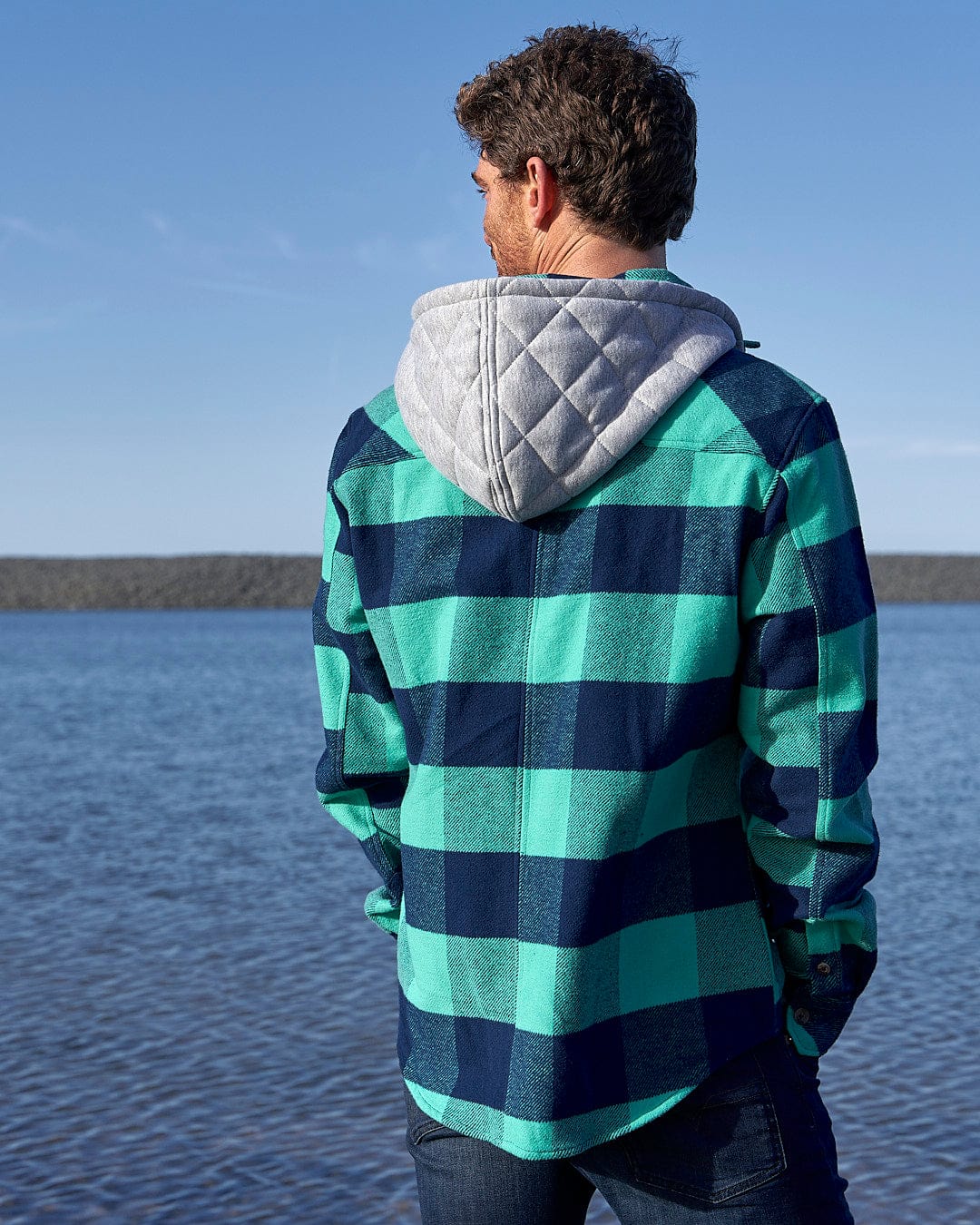 A man wearing a Saltrock Beale - Mens Heavy Weight Hooded Shirt - Dark Green standing by the water.
