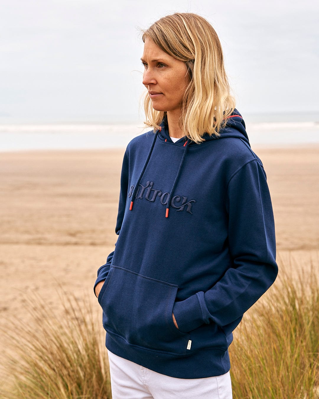 A woman wearing a Saltrock Celeste Pipe - Womens Pop Hoodie in Blue, standing on a beach.