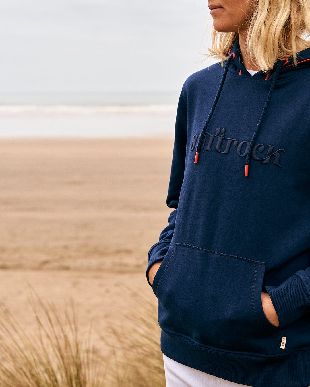 A woman wearing a Saltrock Celeste Pipe - Womens Pop Hoodie - Blue standing on a beach.