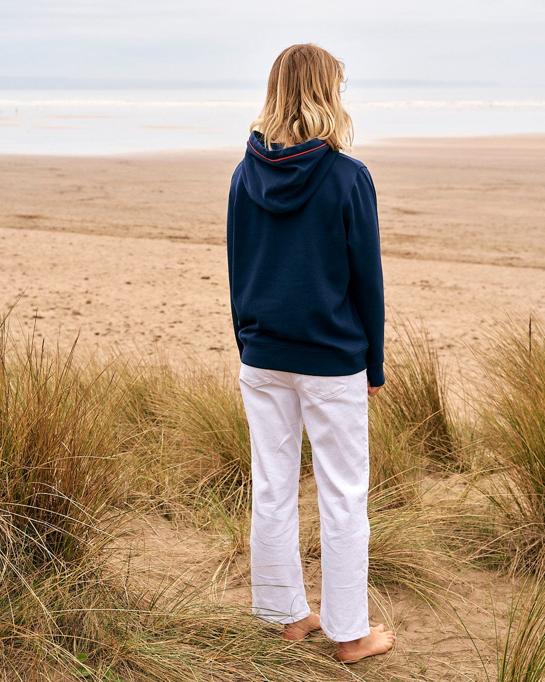 A woman standing on a beach looking at the ocean wearing a Saltrock Celeste Pipe - Womens Pop Hoodie in Blue.