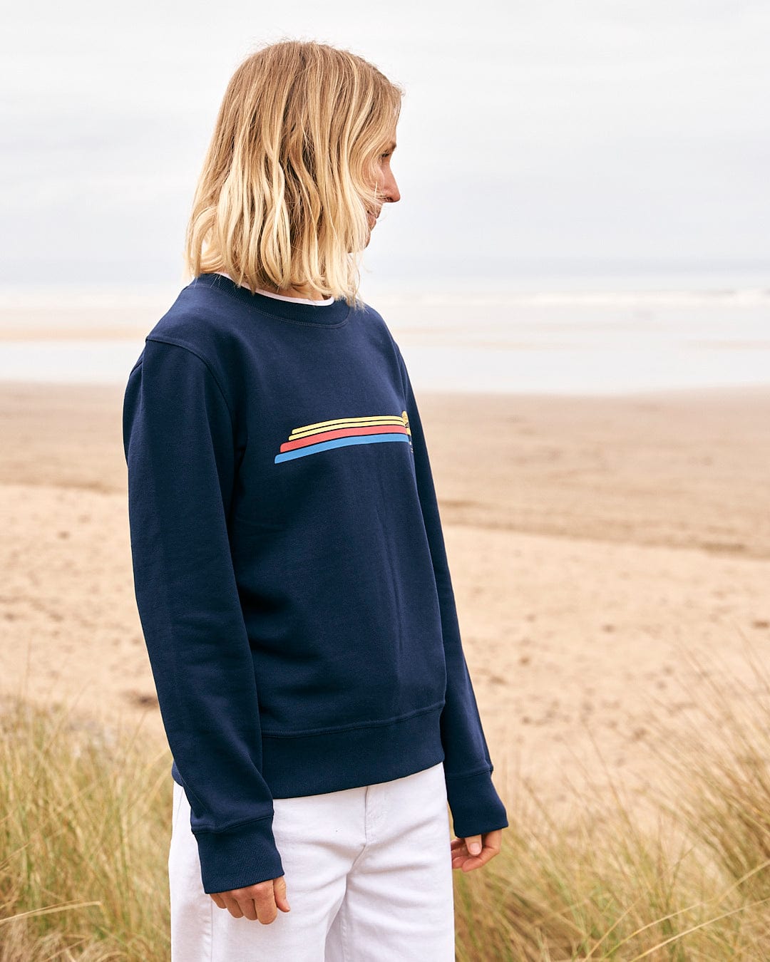 A woman wearing a Celeste Stripe - Womens Crew Sweat - Blue by Saltrock standing on the beach.