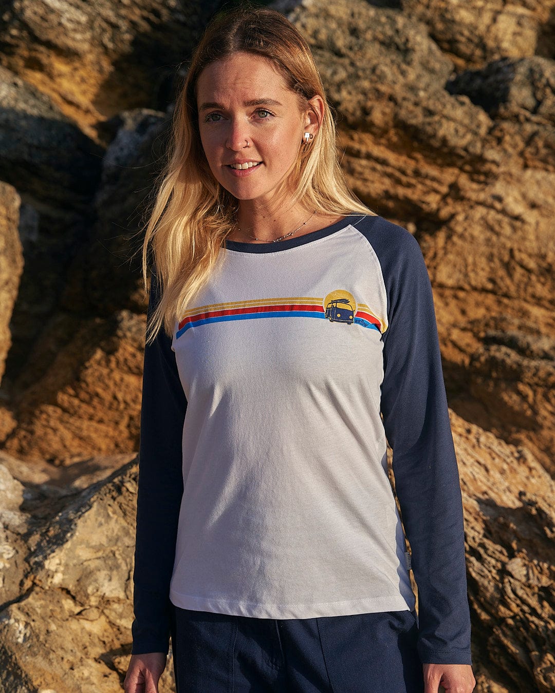 A woman is standing in front of rocks wearing a Celeste Stripe - Womens Long Sleeve Raglan T-Shirt - White/navy from Saltrock.