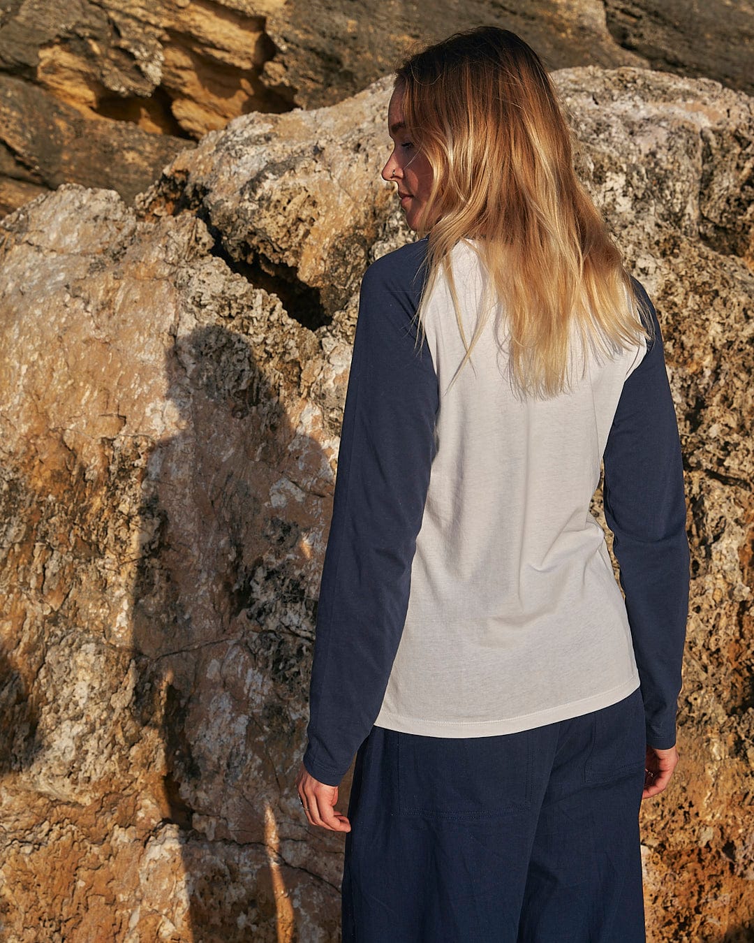 A woman wearing a Saltrock Celeste Stripe - Womens Long Sleeve Raglan T-Shirt - White/navy and pants is standing on a rock.