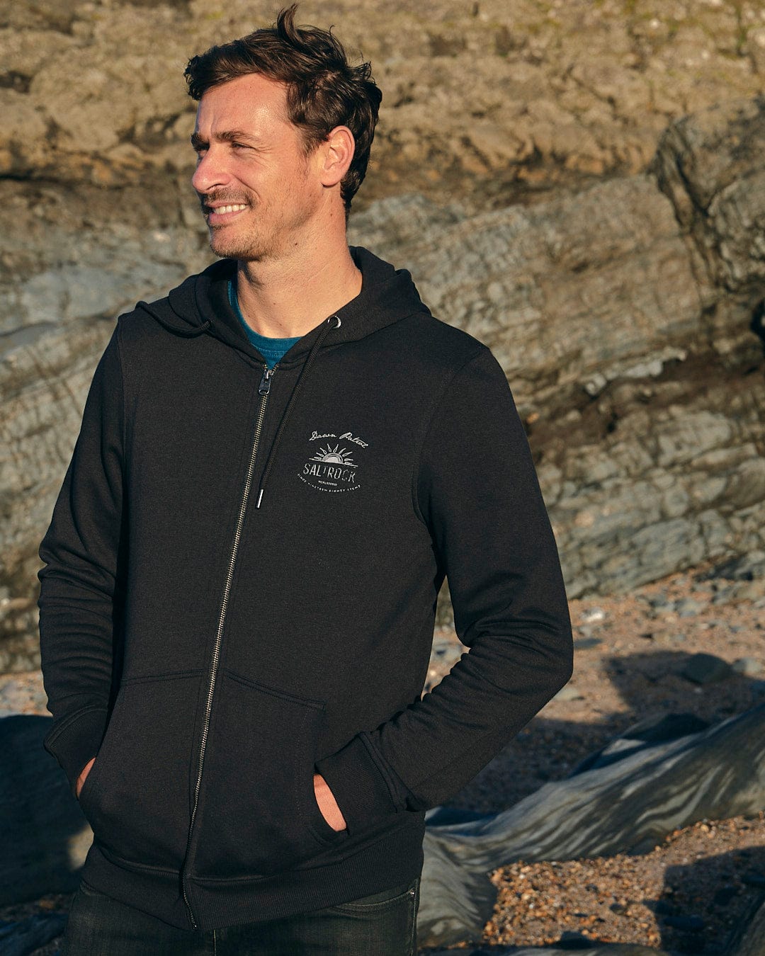 A man wearing a Saltrock Dawn Patrol - Mens Zip Hoodie - Black standing next to rocks.