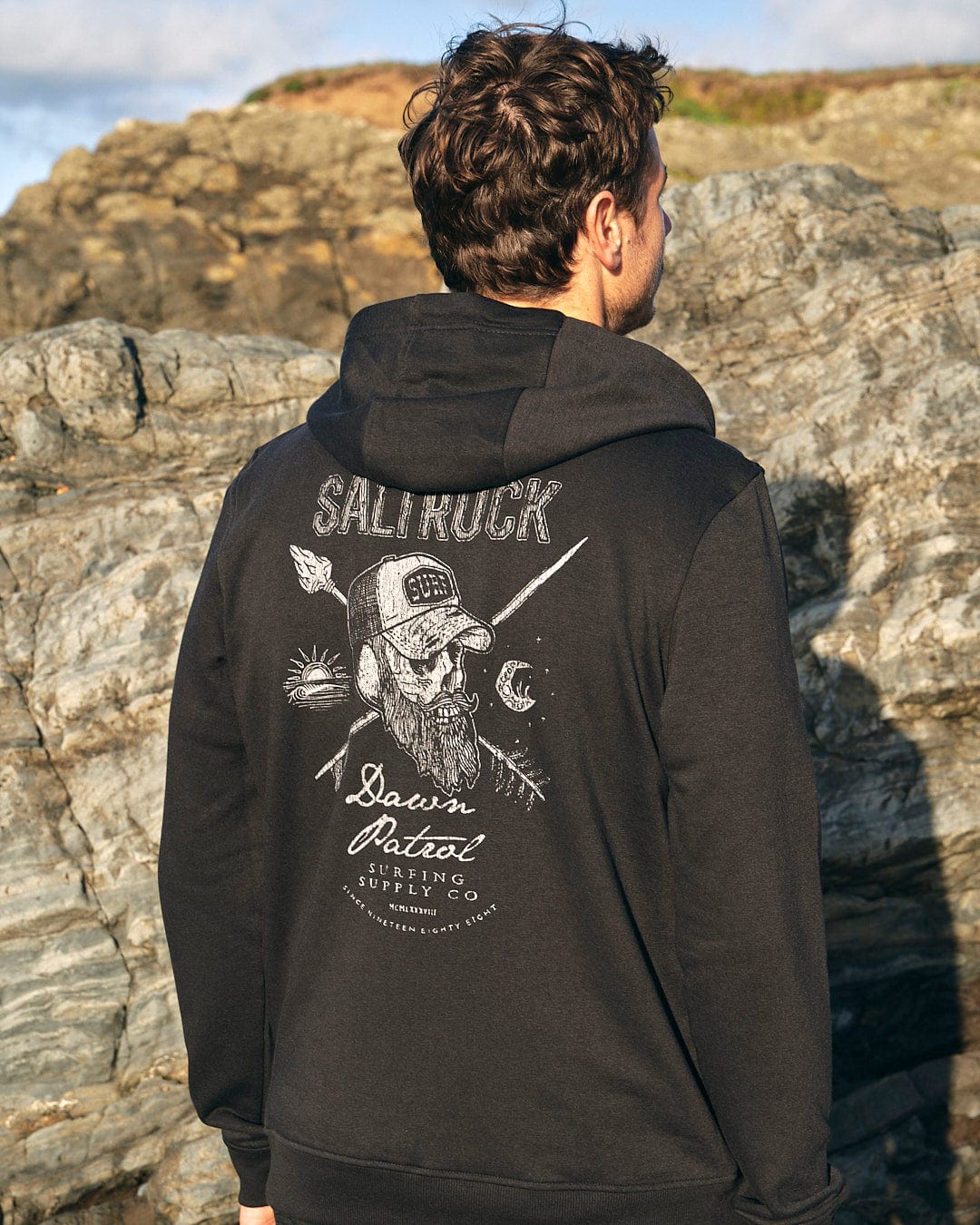 The back of a man wearing a Saltrock Dawn Patrol - Mens Zip Hoodie - Black with a skull and crossbones.