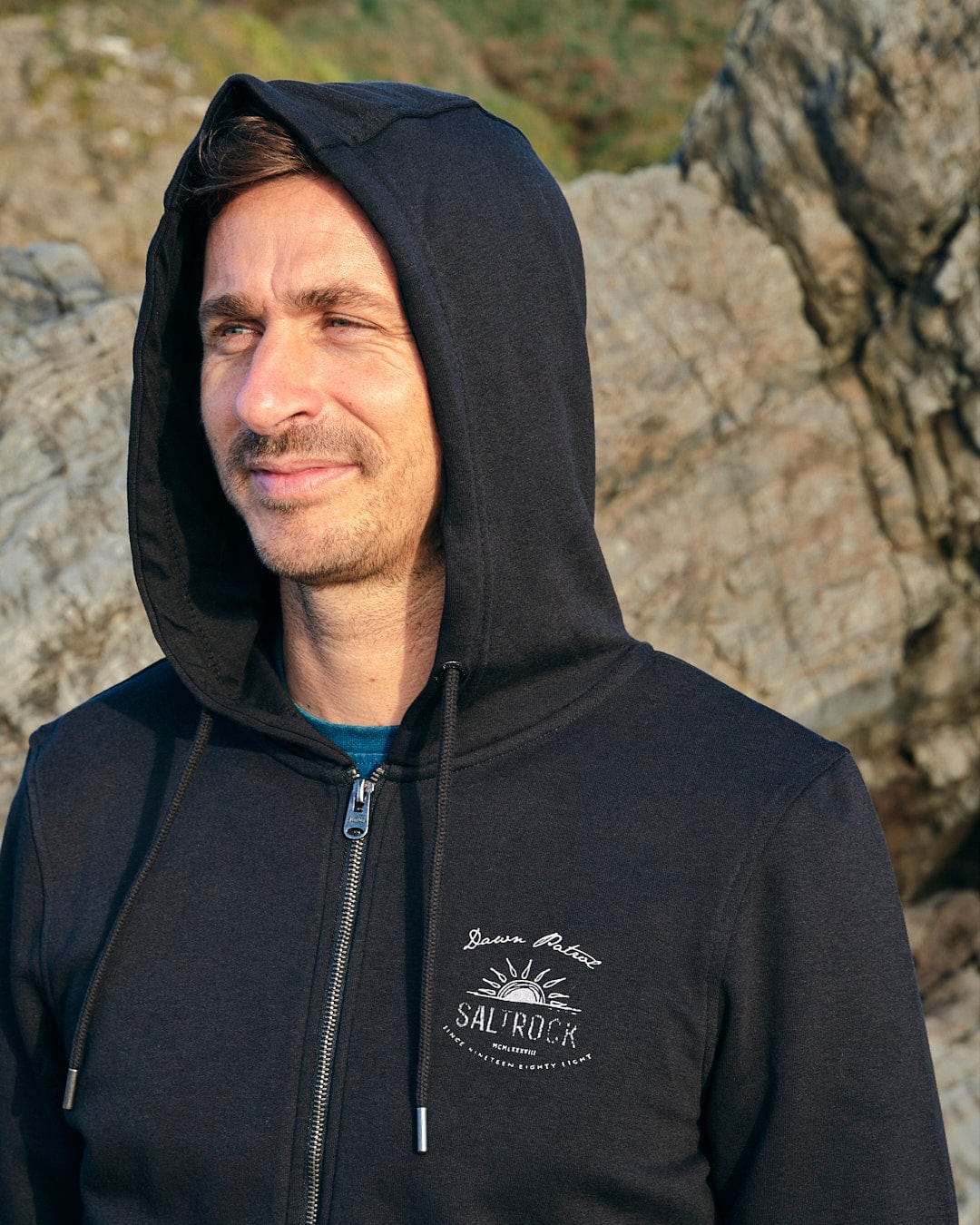 A man wearing a Saltrock Dawn Patrol - Mens Zip Hoodie - Black standing next to rocks.