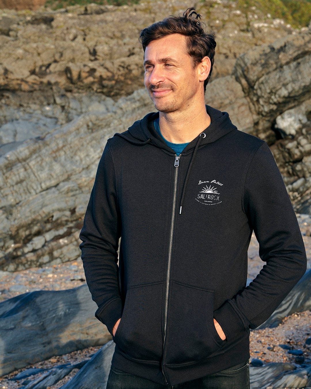 A man wearing a Saltrock Dawn Patrol - Mens Zip Hoodie - Black standing on a rocky beach.