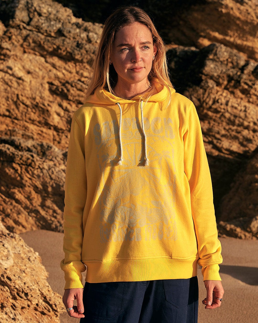 A woman wearing a Saltrock Dreamseeker - Womens Pop Hoodie - Yellow standing on a rocky beach.