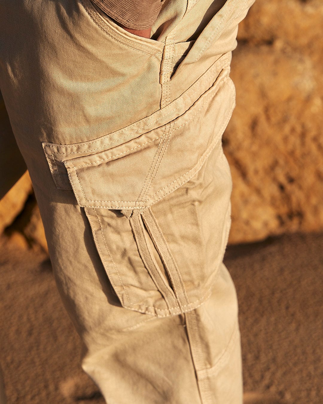 A man wearing Saltrock's Godrevy - Mens Cargo Trouser in Cream is standing on a beach with a cell phone in his pocket.