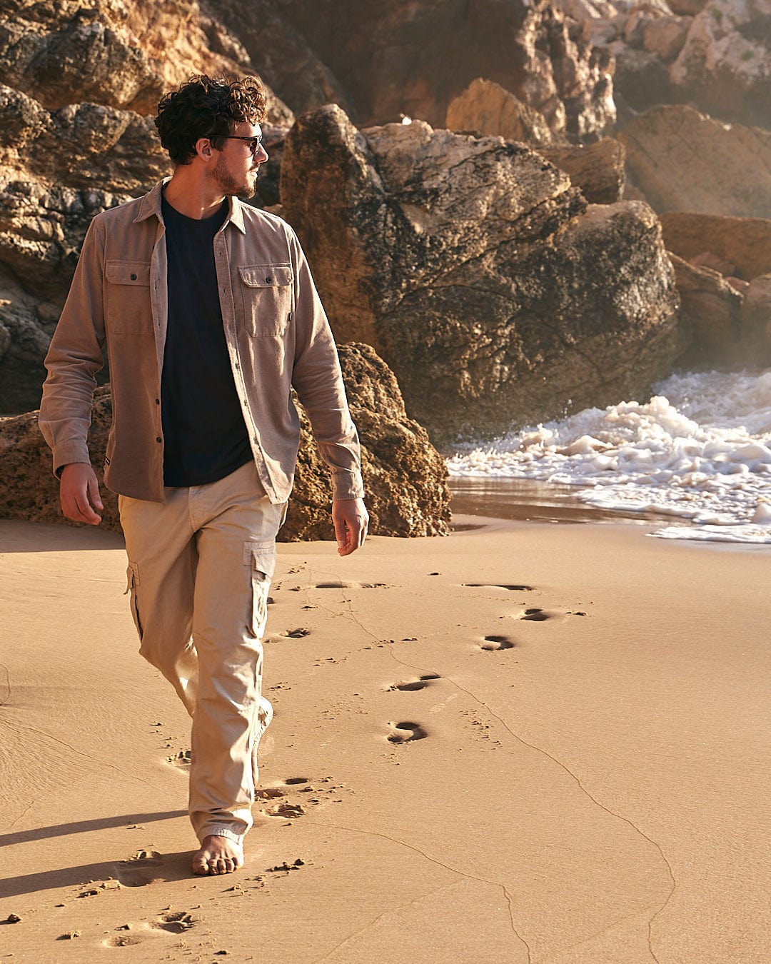A man in Saltrock's Godrevy - Mens Cargo Trouser - Cream walking on a beach with a surfboard, enjoying an Outdoor Adventure by the sea.