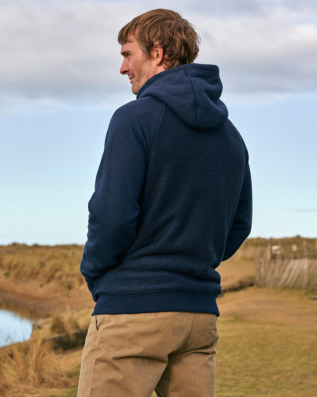 A man in a Saltrock Hall - Mens Orkney Borg Lined Hoodie - Dark Blue standing next to a body of water.
