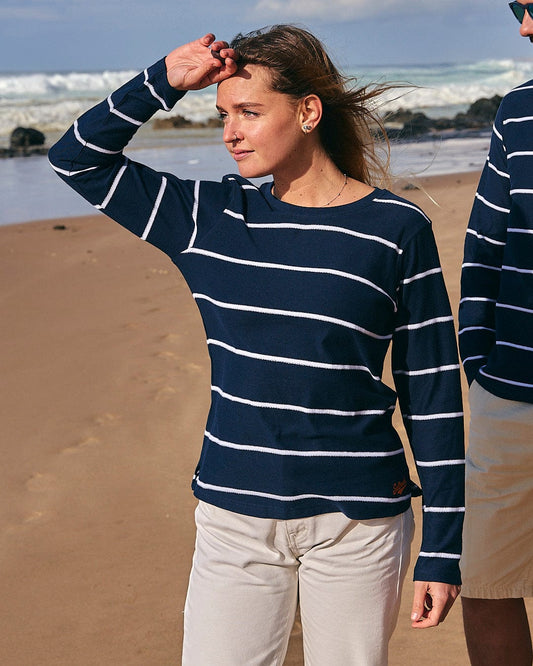 A woman in a Saltrock womens Hartland Striped Long Sleeve T-Shirt - Blue shields her eyes from the sun while standing on a beach, with a portion of a man visible beside her.
