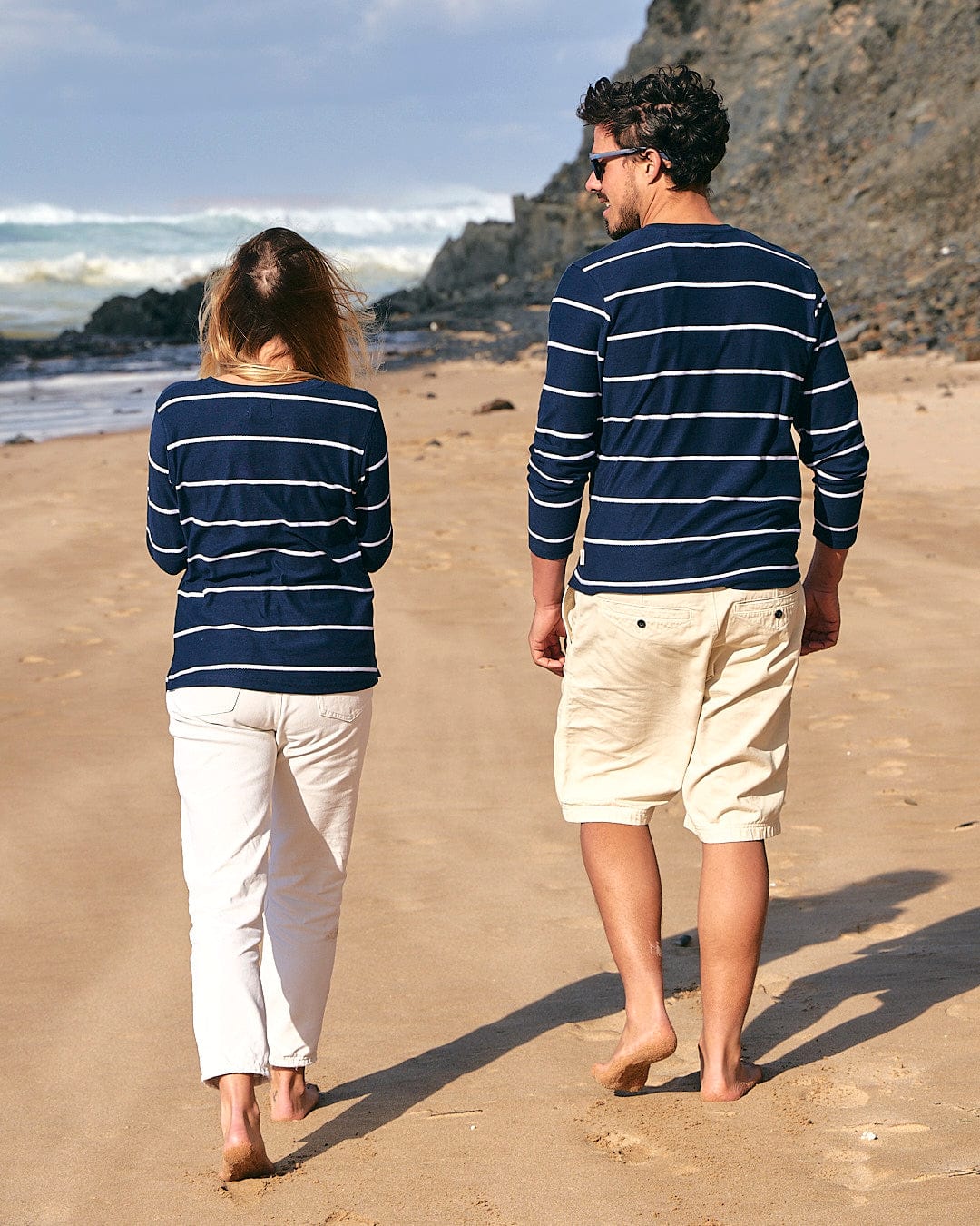 A man and a woman wearing matching Saltrock Hartland Womens Striped Long Sleeve T-Shirts in Blue and beige pants walking on a sandy beach, facing away from the camera, both sporting a nautical look.