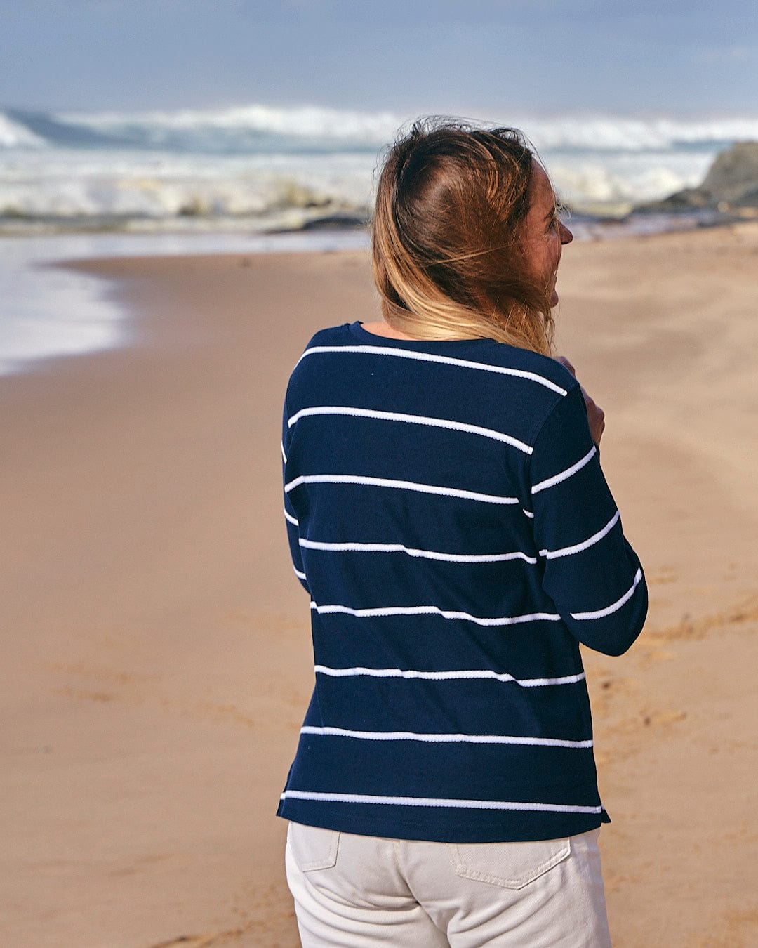 Woman in a Saltrock Hartland - Womens Striped Long Sleeve T-Shirt - Blue looking out at the ocean on a sandy beach.