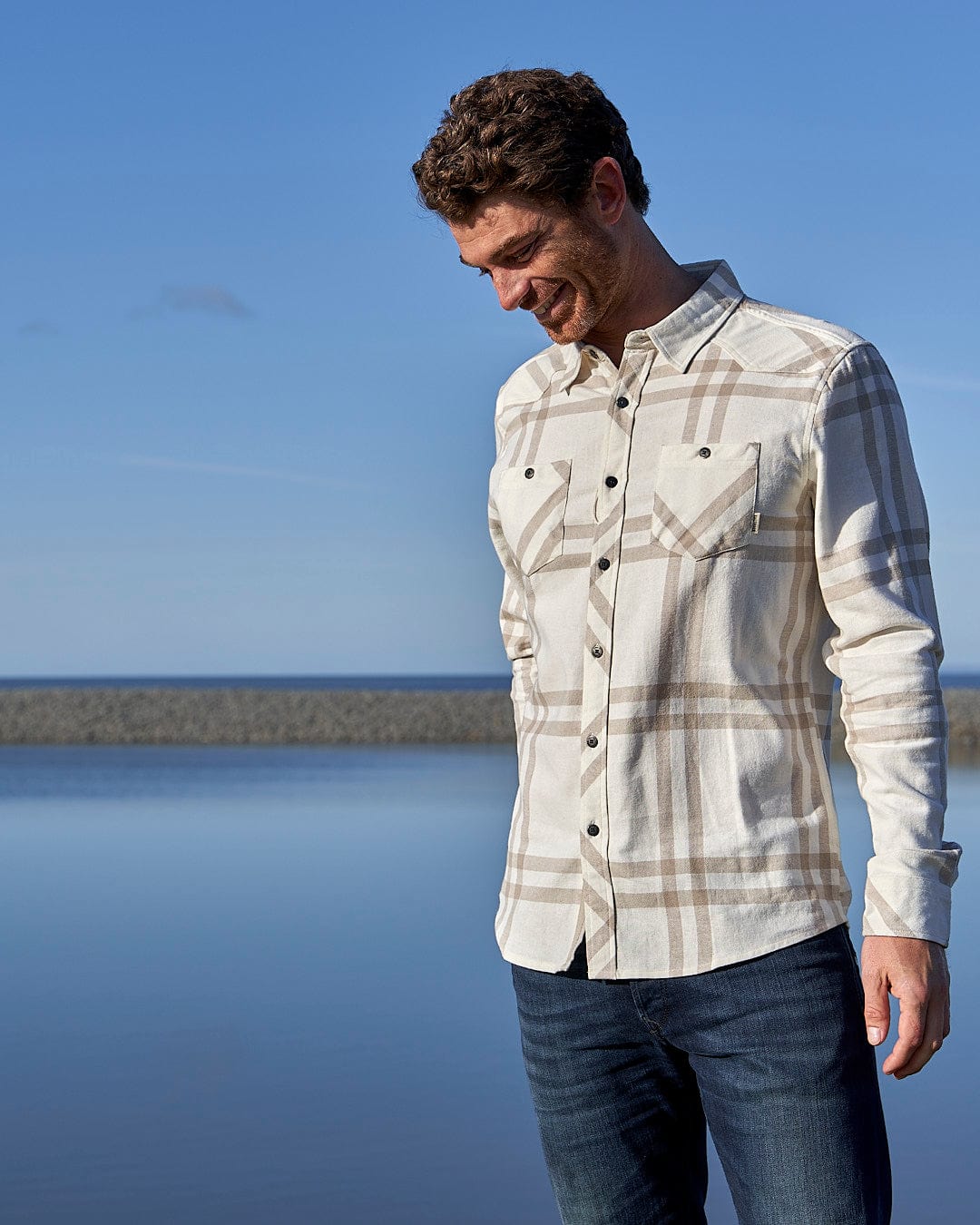 A man in a Saltrock Lazer - Mens Long Sleeve Check Shirt - Light Grey standing by the water.