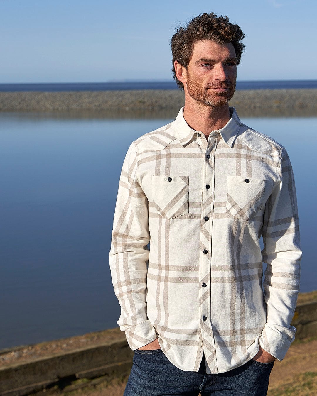 A man is standing next to a body of water wearing a Saltrock Lazer - Mens Long Sleeve Check Shirt - Light Grey.