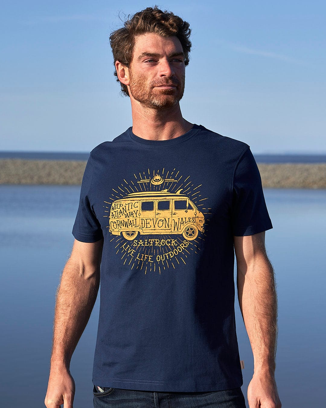 A man wearing a Saltrock Live Life Location - Mens Short Sleeve T-Shirt - Blue standing by the water.