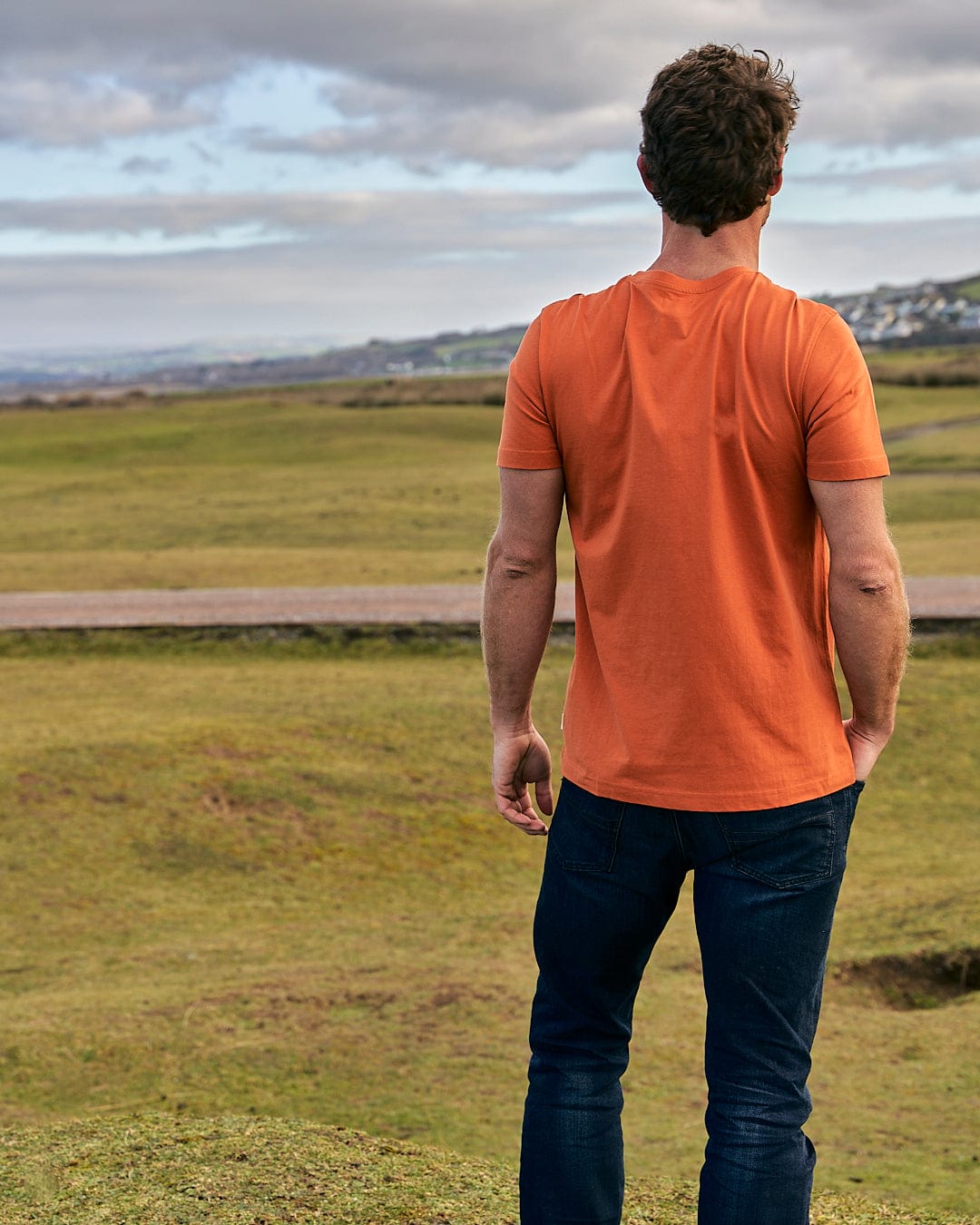 A man in a Saltrock Live Life Location - Mens Short Sleeve T-Shirt - Orange standing on top of a grassy hill.