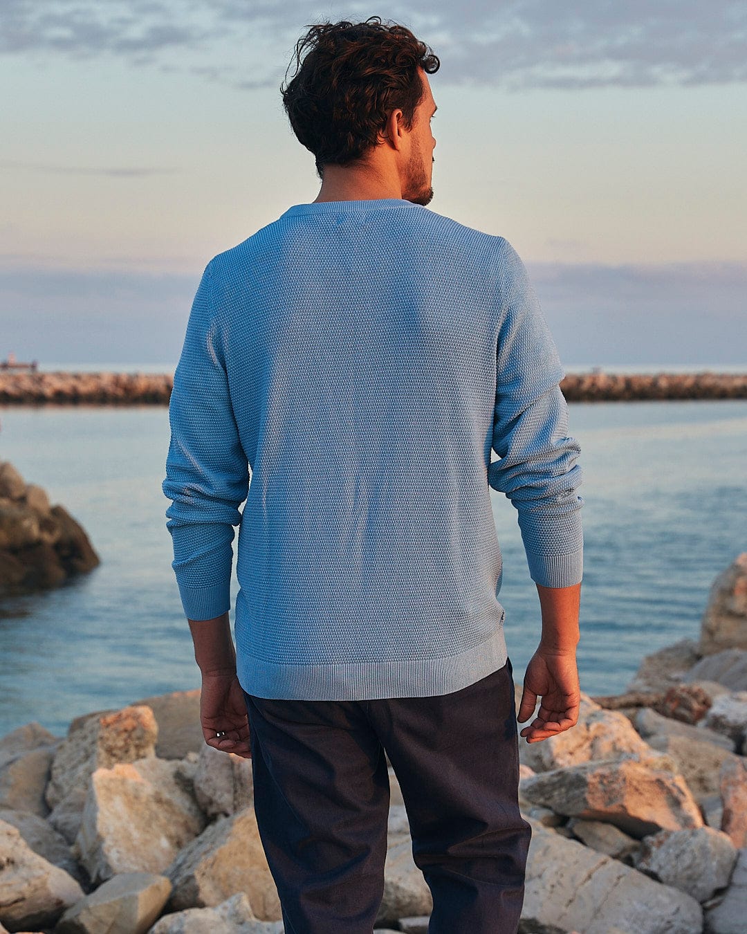 A man wearing the Saltrock Moss - Mens Washed Knitted Crew - Light Blue sweater is standing on rocks near the ocean.