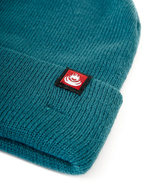 A Saltrock Tight Knit Beanie in Teal with a red logo, perfect for staying cozy in the winter.