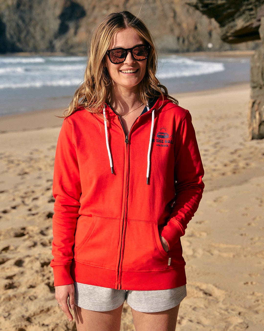 A woman is standing on the beach wearing a Saltrock On The Road - Womens Zip Hoodie - Red.