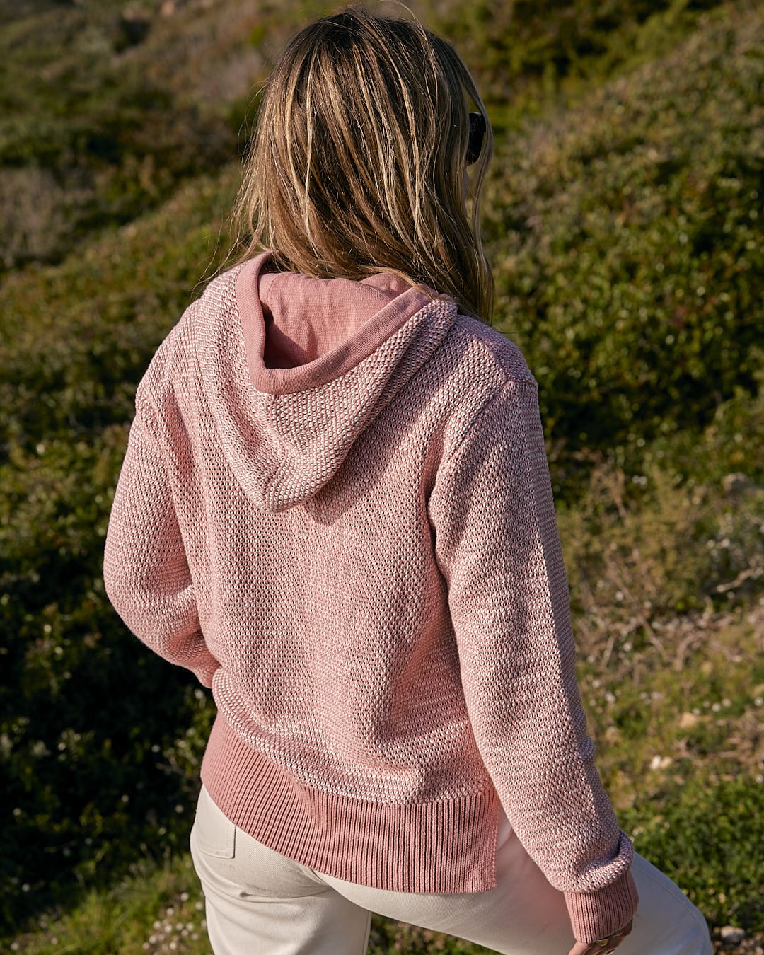 A woman wearing a Saltrock Poppy - Womens Knitted Pop Hoodie - Mid Pink hoodie and white pants.