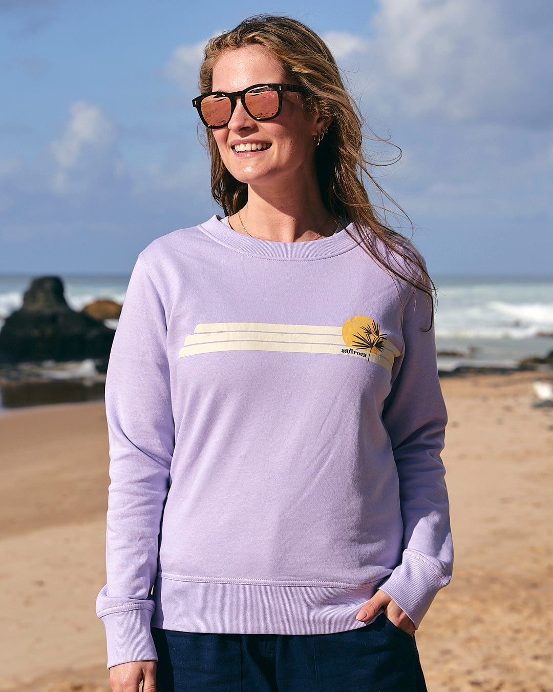 A woman wearing Saltrock sunglasses and a Saltrock Poster Stripe - Womens Crew Sweat - Light Purple on the beach.