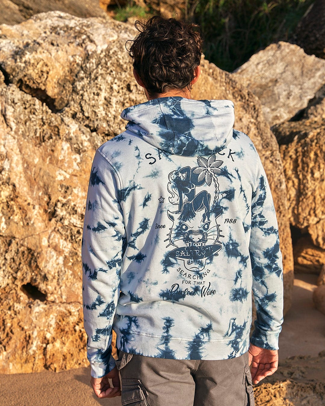 A man wearing a Saltrock Purfect Wave - Mens Tie Dye Pop Hoodie - Light Blue standing on rocks.
