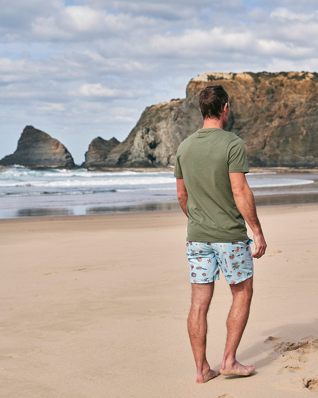 A man standing on a beach looking at the ocean wearing Saltrock - Ruan Mens Swimshorts - Light Blue.