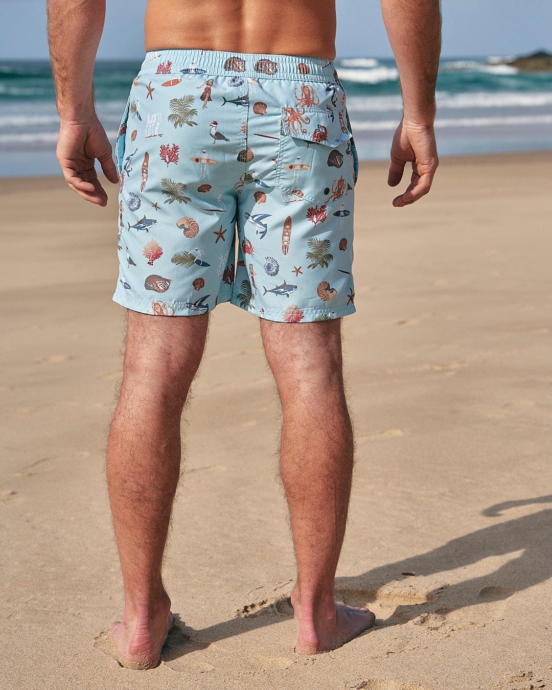 The man standing on the beach is wearing the Saltrock Ruan - Mens Swimshorts - Light Blue.