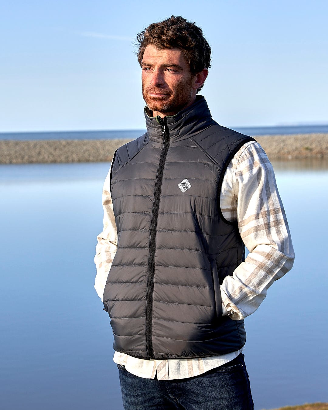 A man standing next to a body of water wearing a Saltrock - Mens Reversible Gilet - Dark Green/Black.
