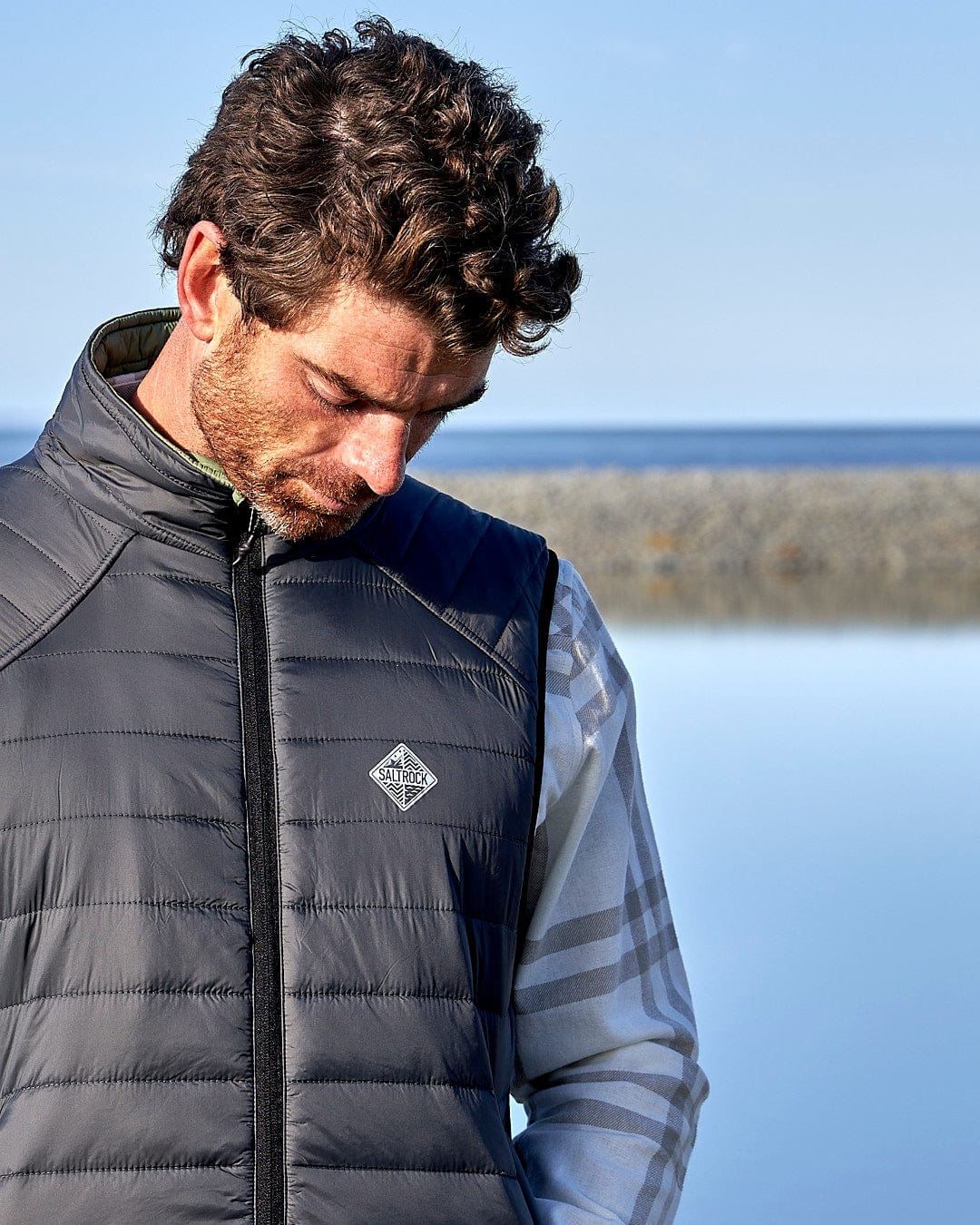 A man is standing by the water wearing a Saltrock - Mens Reversible Gilet - Dark Green/Black.