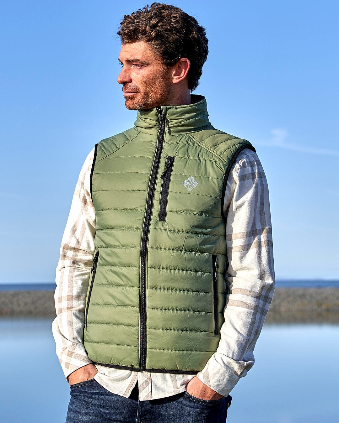 A man in a Saltrock - Scott Mens Reversible Gilet - Dark Green/Black vest, standing by the water.