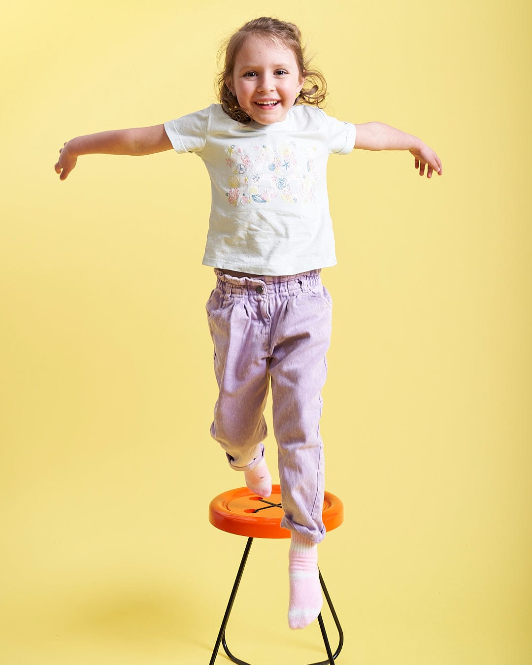 A girl standing on a Saltrock Seabed - Kids Short Sleeve T-Shirt - Light Blue stool.