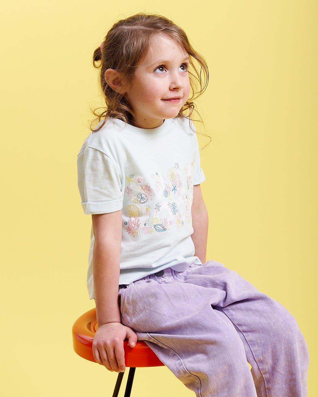 A little girl sitting on a stool wearing a Saltrock Seabed - Kids Short Sleeve T-Shirt - Light Blue.