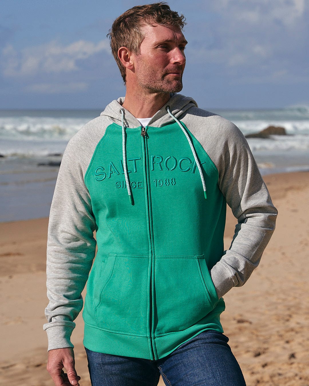 A man standing on a beach wearing a Saltrock Stencil - Mens Zip Hoodie - Green.